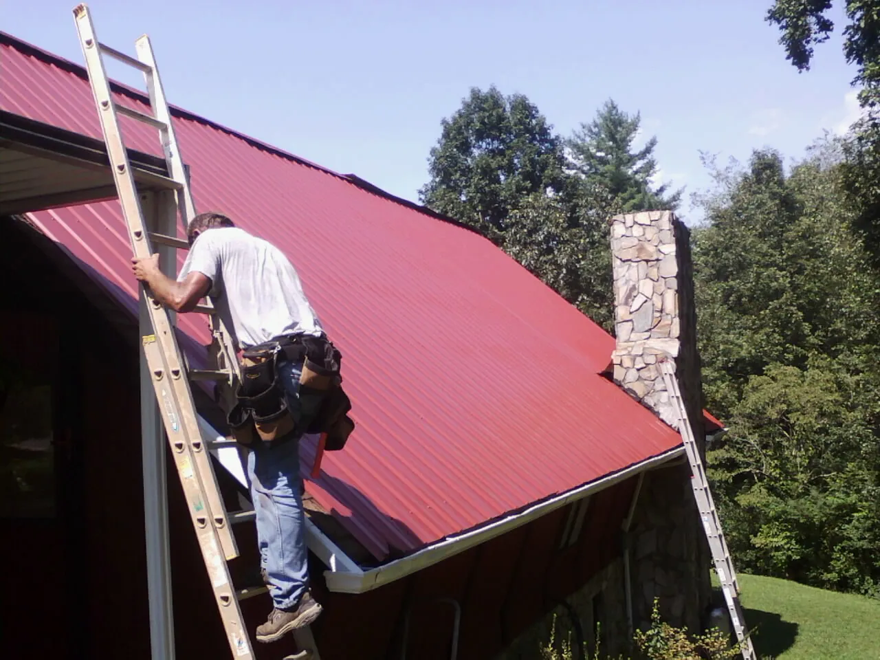 Roofer on a ladder beside a red metal roof and stone chimney — Soffit & Fascia Repair by Roofing Brothers in Coulter